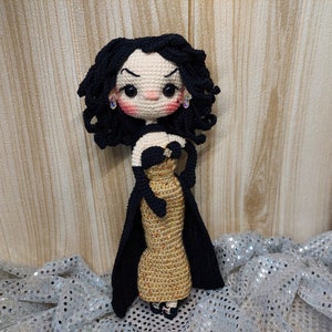 May include: A crocheted doll wearing a black dress with a gold sequined bodice and a black cape. The doll has black hair, brown eyes, and rosy cheeks.