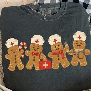 Puede incluir: Sudadera de cuello redondo gris oscuro con un gráfico de cuatro hombres de jengibre vestidos de enfermeras. Cada hombre de jengibre lleva un gorro de enfermera blanco con una cruz roja. Uno sostiene un bastón de caramelo, otro un maletín médico y otro una jeringa.