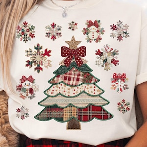 Op de afbeelding: Crèmekleurig T-shirt met een kerstboomontwerp gemaakt van ruitpatronen, strikken en een ster. Sneeuwvlokken en een sneeuwpop maken ook deel uit van het feestelijke ontwerp. Een casual, feestelijk kledingstuk.