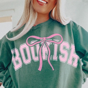 May include: A green sweatshirt with the word "BOUSKISH" printed in white with a pink bow on top of the word.