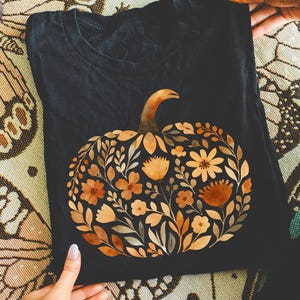 Może przedstawiać: Czarny t-shirt z kwiatowym wzorem dyni w odcieniach pomarańczu, brązu i ż&oacute;łci. Dynia składa się z r&oacute;żnych kwiat&oacute;w i liści. Koszulka jest trzymana przez osobę.