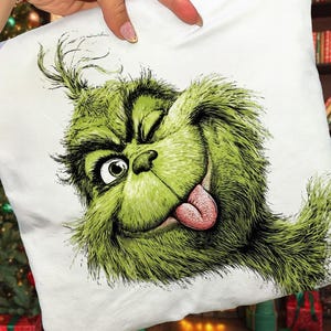 May include: White square pillow featuring a green cartoon character with a winking eye and tongue sticking out. The character has green fur and a mischievous expression. The pillow is held up by a hand.