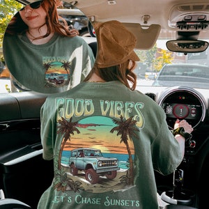 Op de afbeelding: Een groen T-shirt met een afbeelding van een vintage blauwe jeep die op een strand bij zonsondergang rijdt. De tekst "Good Vibes Let's Chase Sunsets" is op het shirt gedrukt.