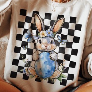 May include: A white sweatshirt with a black and white checkered pattern and a blue bunny holding a blue Easter egg. The bunny is wearing a flower crown and there are butterflies around it.