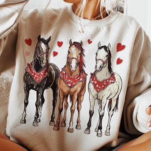 Puede incluir: Una sudadera blanca con un gr&aacute;fico de tres caballos con bandanas rojas y rodeados de corazones rojos. La sudadera la lleva puesta una persona sentada en un sof&aacute;.