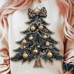 May include: A cream-colored sweatshirt featuring a Christmas tree design. The tree is decorated with gold ornaments, a leopard print bow, and a star. The tree is embellished with gold glitter and a gold bell at the base. The sweatshirt is a festive holiday apparel item.