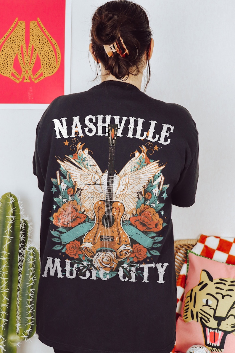 May include: Black t-shirt with a graphic print of a guitar, wings, and flowers. The text "Nashville Music City" is printed on the shirt.