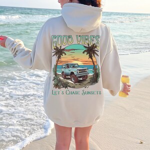 Op de afbeelding: Een cr&egrave;mekleurige hoodie met een grafische print van een vintage terreinwagen die op een strand rijdt met palmbomen en een zonsondergang. De tekst "GOOD VIBES" en "LET'S CHASE SUNSETS" staat op de hoodie gedrukt.