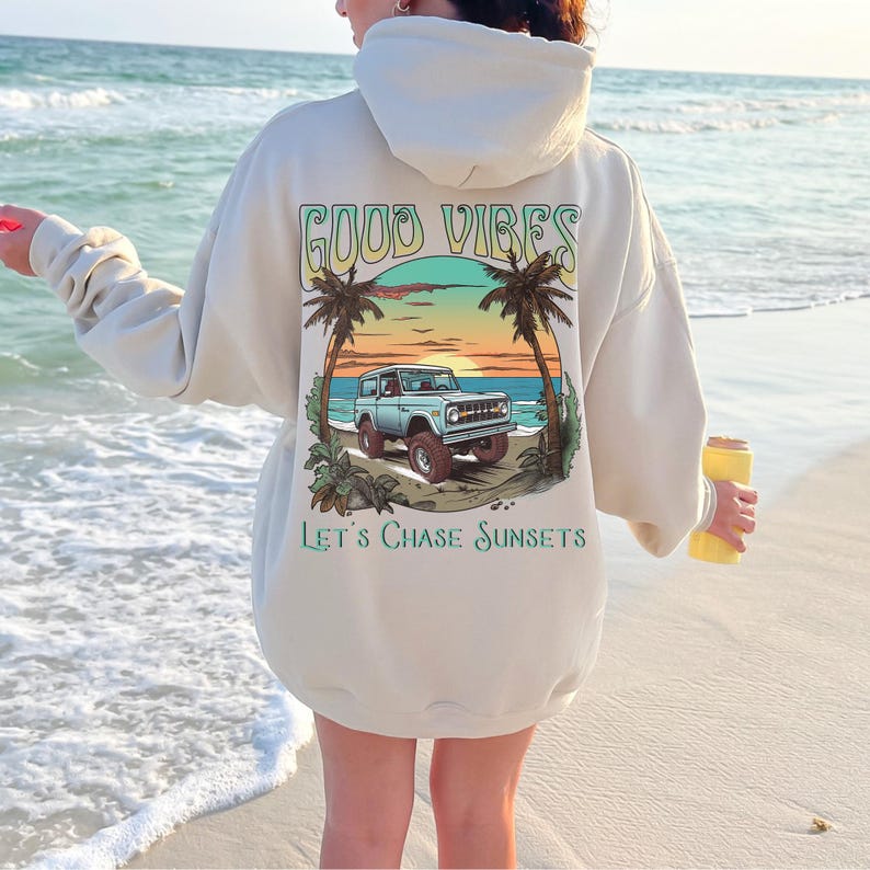 Op de afbeelding: Een cr&egrave;mekleurige hoodie met een grafische print van een vintage terreinwagen die op een strand rijdt met palmbomen en een zonsondergang. De tekst "GOOD VIBES" en "LET'S CHASE SUNSETS" staat op de hoodie gedrukt.