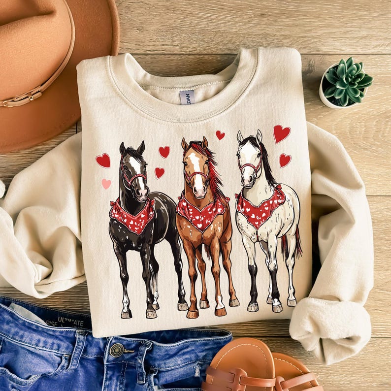 Puede incluir: Una sudadera beige con un gr&aacute;fico de tres caballos con bandanas rojas con corazones. Los caballos son marr&oacute;n, negro y blanco.