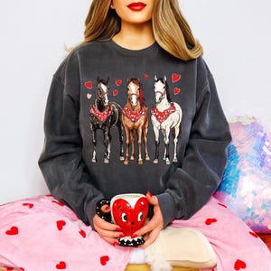 Puede incluir: Una sudadera gris oscuro con un gr&aacute;fico de tres caballos con pa&ntilde;uelos y corazones. La sudadera la lleva puesta una persona que lleva un pijama rosa con corazones.