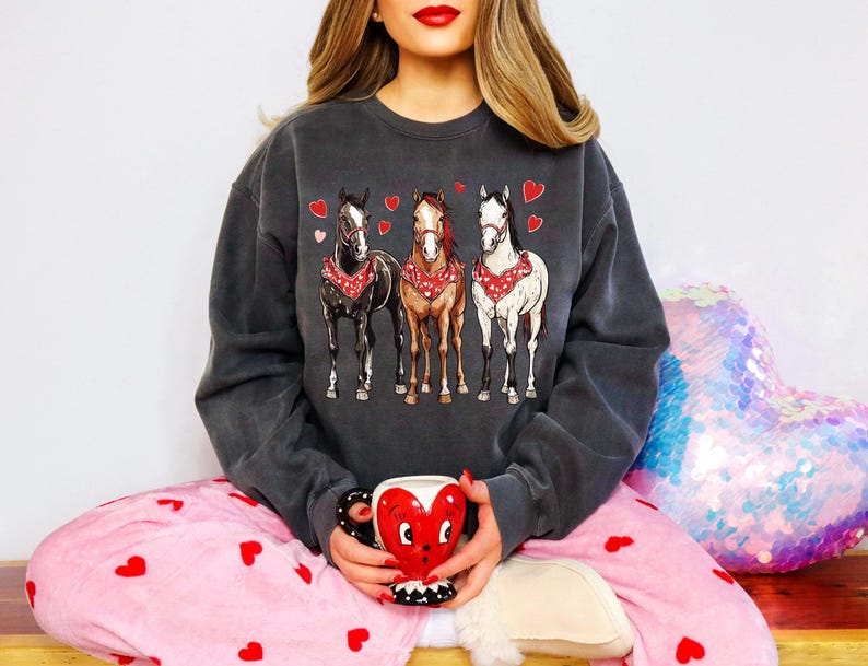 Puede incluir: Una sudadera gris oscuro con un gr&aacute;fico de tres caballos con pa&ntilde;uelos y corazones. La sudadera la lleva puesta una persona que lleva un pijama rosa con corazones.