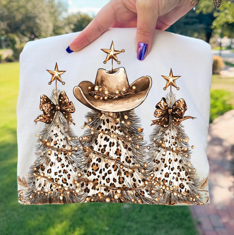 May include: White sweatshirt featuring a Christmas tree design with a cowboy hat, leopard print, and gold star accents. The trees are adorned with string lights and bows. A festive and unique holiday apparel item.