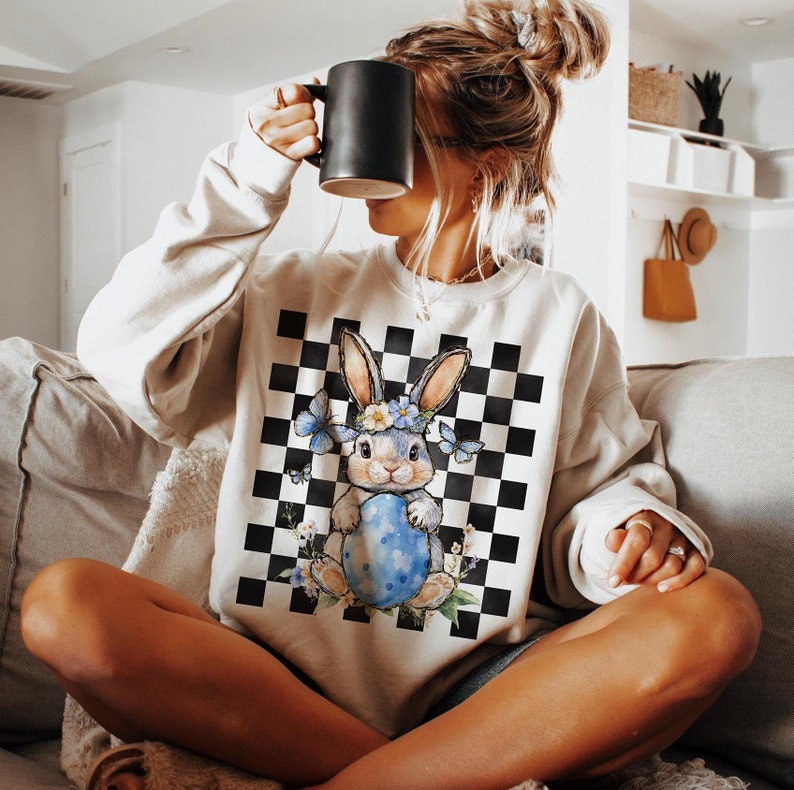 May include: A white sweatshirt with a black and white checkered pattern and a blue bunny holding a blue Easter egg. The bunny is wearing a flower crown and there are butterflies around it.