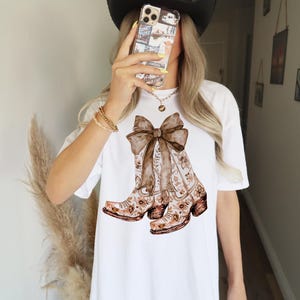 Puede incluir: Camiseta blanca con un estampado floral marr&oacute;n de botas de vaquero atadas con un lazo marr&oacute;n.