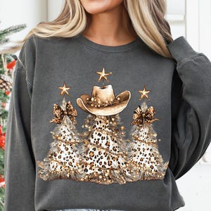 May include: Dark gray sweatshirt featuring a festive design. The graphic includes three Christmas trees with leopard print, adorned with a cowboy hat, gold stars, and string lights. The design has a country and holiday theme.