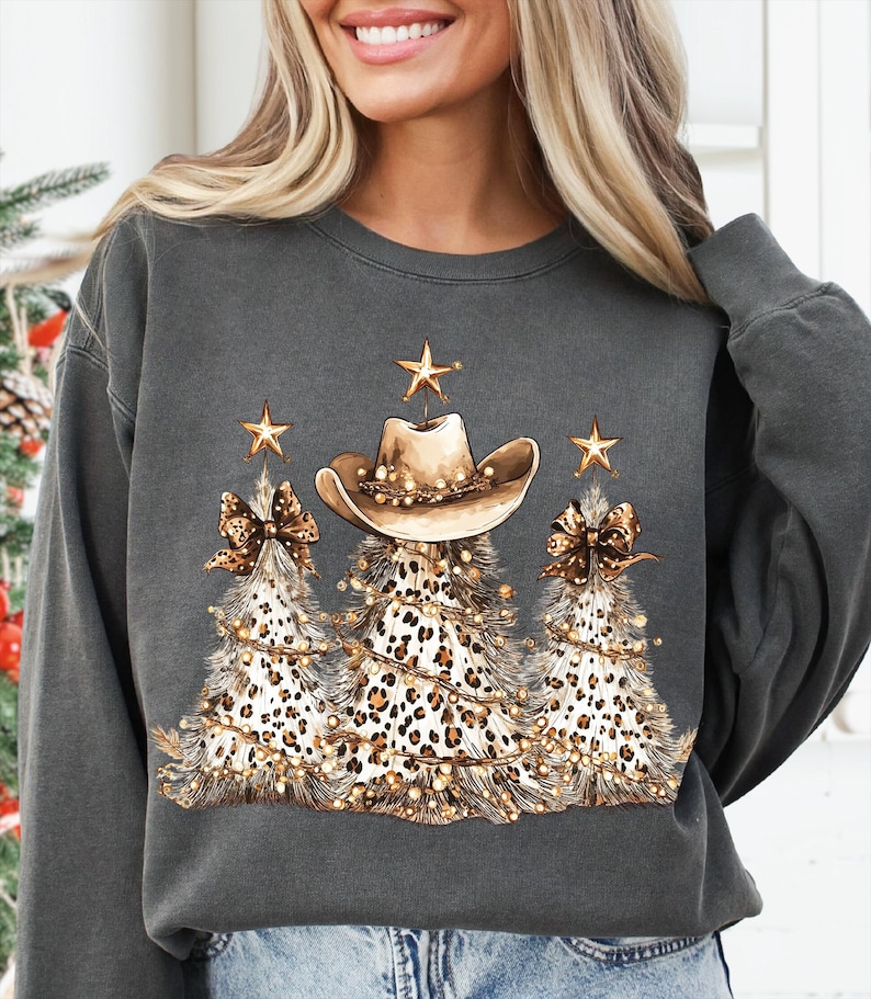 May include: Dark gray sweatshirt featuring a festive design. The graphic includes three Christmas trees with leopard print, adorned with a cowboy hat, gold stars, and string lights. The design has a country and holiday theme.