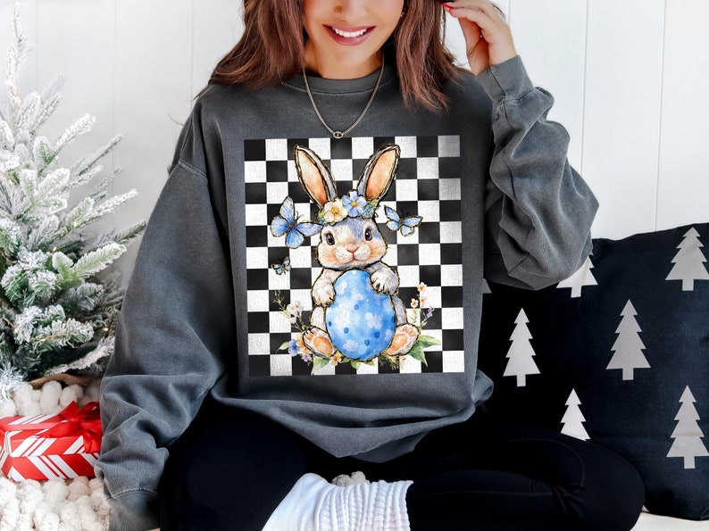 May include: A gray sweatshirt with a checkered pattern and a cartoon bunny holding a blue Easter egg. The bunny is wearing a flower crown and blue butterfly wings.