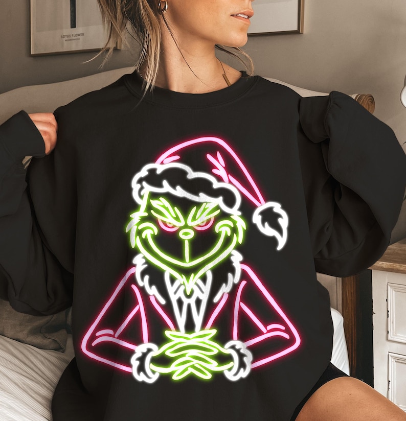 Puede incluir: Sudadera negra con un dise&ntilde;o de ne&oacute;n de un personaje verde con gorro de Pap&aacute; Noel y traje rojo. El dise&ntilde;o tiene contornos blancos y acentos rosas y verdes, creando un tema festivo navide&ntilde;o.