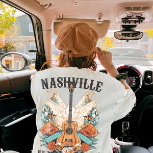 May include: A white t-shirt with a graphic design featuring a guitar, wings, and flowers. The text "Nashville Music City" is printed on the shirt.