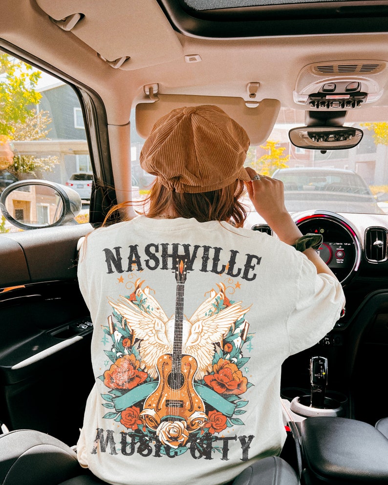 May include: A white t-shirt with a graphic design featuring a guitar, wings, and flowers. The text "Nashville Music City" is printed on the shirt.