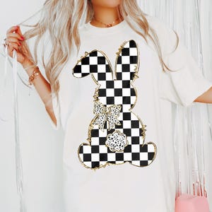 May include: White t-shirt with a black and white checkered bunny silhouette with a gold glitter outline. The bunny has a black and white polka dot bow.