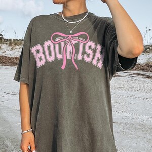 May include: A gray oversized t-shirt with a pink bow and the word "BOUSH" printed on the front. The shirt is made of a soft, comfortable material and has a relaxed fit.