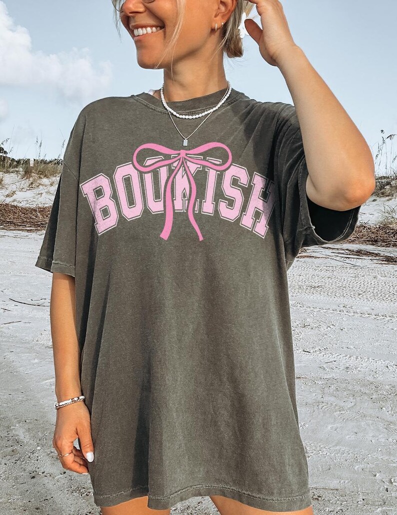 May include: A gray oversized t-shirt with a pink bow and the word "BOUSH" printed on the front. The shirt is made of a soft, comfortable material and has a relaxed fit.