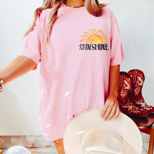 May include: A pink oversized t-shirt with a graphic of a yellow sun and the word "Sunshine" in black text.