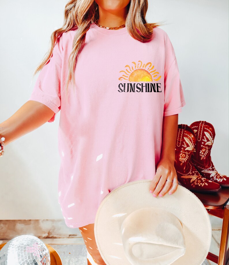 May include: A pink oversized t-shirt with a graphic of a yellow sun and the word "Sunshine" in black text.