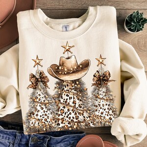 May include: Cream-colored sweatshirt featuring a festive design. The graphic includes three Christmas trees with a leopard print pattern, cowboy hats, bows, and star ornaments. The trees are adorned with string lights.