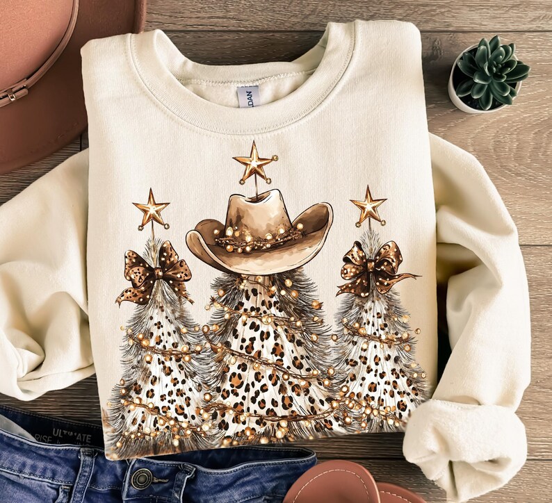 May include: Cream-colored sweatshirt featuring a festive design. The graphic includes three Christmas trees with a leopard print pattern, cowboy hats, bows, and star ornaments. The trees are adorned with string lights.