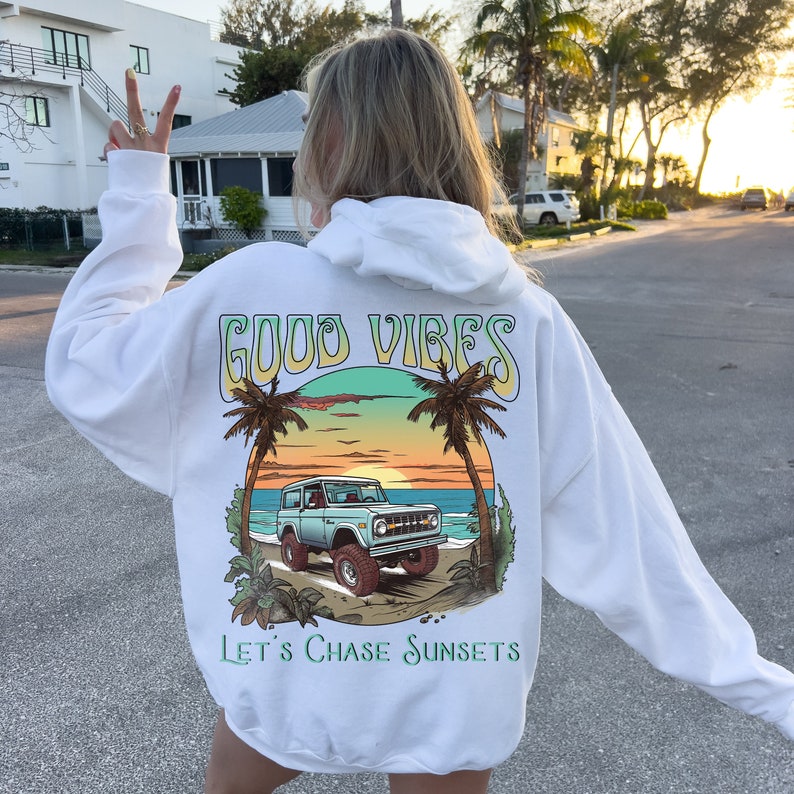 Op de afbeelding: Witte hoodie met een grafische print van een vintage auto die bij zonsondergang over een strand rijdt. De grafiek bevat ook palmbomen en de tekst "GOOD VIBES" en "LET'S CHASE SUNSETS".