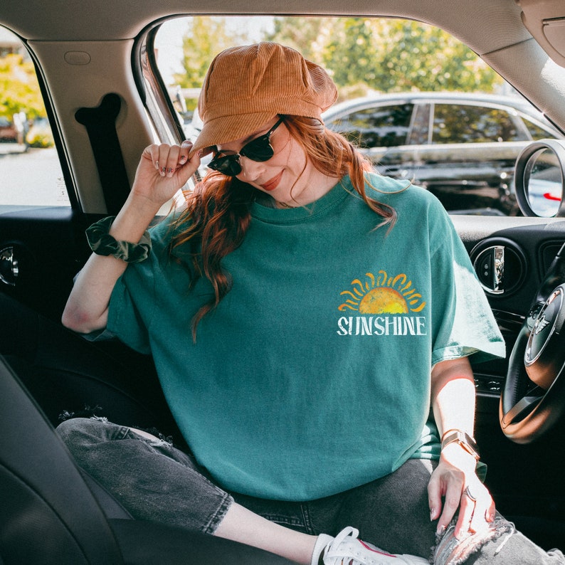 May include: A teal blue t-shirt with a yellow sun graphic and the text "Sunshine" in white. The person is wearing a brown corduroy newsboy cap and sunglasses.