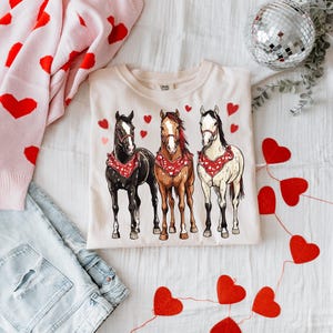 Puede incluir: Una camiseta de color crema con un gr&aacute;fico de tres caballos con bandanas rojas. Los caballos son marr&oacute;n, negro y blanco. El fondo es blanco con corazones rojos.