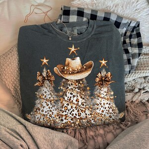 May include: Dark gray sweatshirt featuring a cowboy hat and three Christmas trees with leopard print and string lights. The trees have bows and star accents. A black and white plaid shirt is folded in the background.