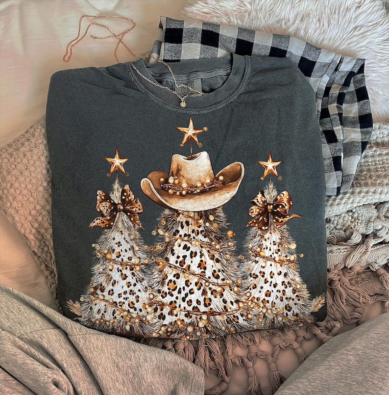 May include: Dark gray sweatshirt featuring a cowboy hat and three Christmas trees with leopard print and string lights. The trees have bows and star accents. A black and white plaid shirt is folded in the background.