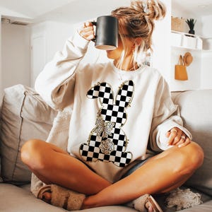 May include: A white sweatshirt with a black and white checkered bunny graphic. The bunny has a gold outline and a gold bow.