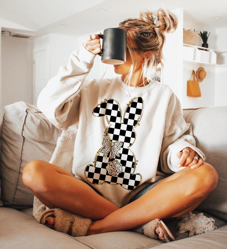 May include: A white sweatshirt with a black and white checkered bunny graphic. The bunny has a gold outline and a gold bow.