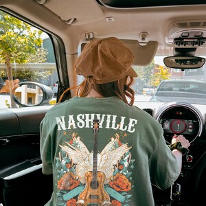 May include: A person wearing a green t-shirt with a graphic of a guitar, wings, and flowers. The shirt says "Nashville Music City".