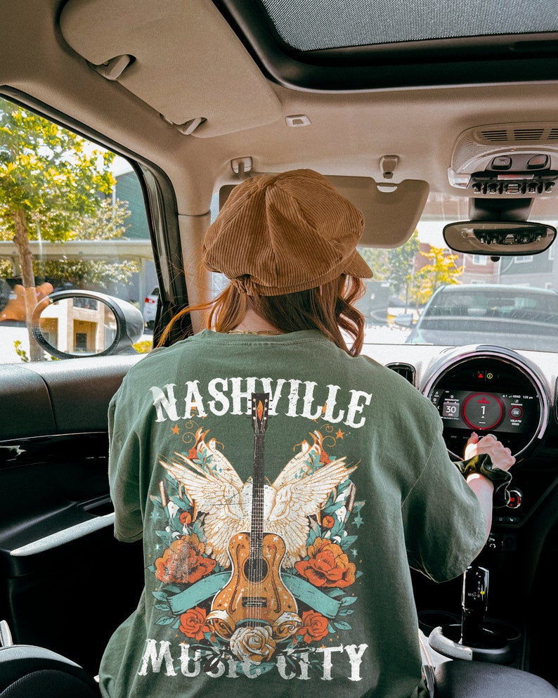 May include: A person wearing a green t-shirt with a graphic of a guitar, wings, and flowers. The shirt says "Nashville Music City".