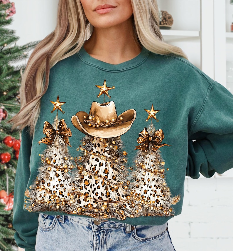 May include: Teal green sweatshirt featuring a festive design. The graphic includes three Christmas trees with leopard print, cowboy hats, and gold star accents. The trees are adorned with string lights and bow accents.