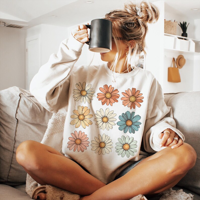 May include: A white sweatshirt featuring a colourful daisy print. The daisies are arranged in a grid pattern and are in shades of pink, yellow, blue, and green.