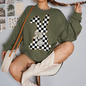 May include: A green sweatshirt with a checkered bunny graphic. The bunny has a gold bow and is outlined in gold. The sweatshirt has long sleeves and a crew neck.