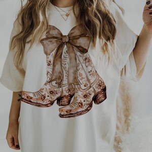 Puede incluir: Camiseta blanca con un estampado floral marr&oacute;n de botas de vaquero atadas con un gran lazo marr&oacute;n.