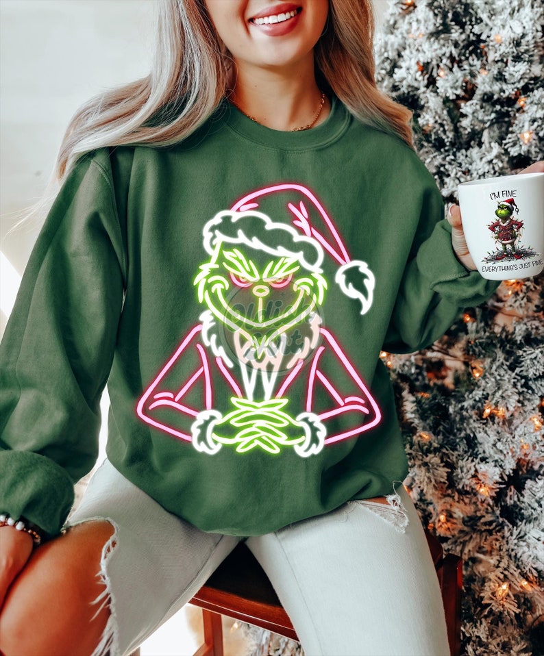 Puede incluir: Sudadera verde oscuro con un dise&ntilde;o de ne&oacute;n del Grinch con un gorro de Pap&aacute; Noel. El Grinch est&aacute; delineado en verde ne&oacute;n y rosa. La persona sostiene una taza blanca con un gr&aacute;fico del Grinch y el texto "I'M FINE. EVERYTHING'S JUST FINE."