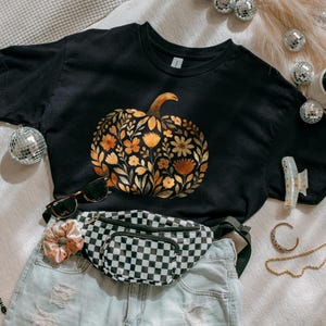 Może przedstawiać: Czarny t-shirt z kwiatowym wzorem dyni w odcieniach pomarańczy, ż&oacute;łci i złota. Koszulka jest stylizowana z czarno-białą nerką w szachownicę, dżinsowymi szortami, okularami przeciwsłonecznymi i akcesoriami. Idealny na jesień.