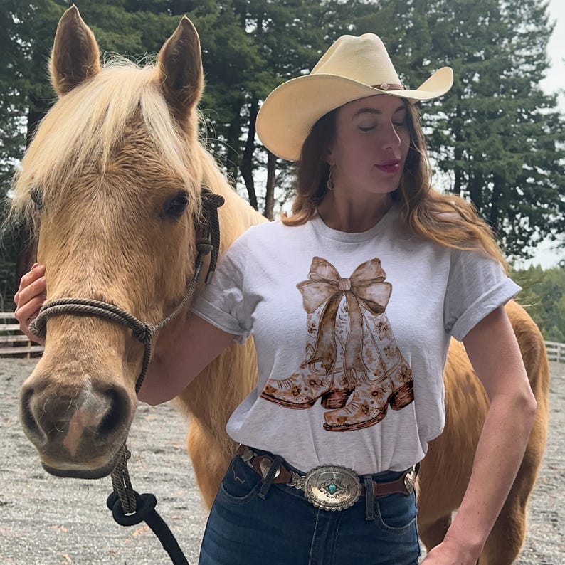 Puede incluir: Una camiseta blanca con un estampado floral marr&oacute;n y blanco de botas de vaquero con un gran lazo. La camiseta la lleva puesta una persona con el pelo largo y casta&ntilde;o y un sombrero de vaquero de paja. La persona est&aacute; de pie junto a un caballo marr&oacute;n claro.