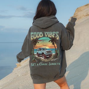Op de afbeelding: Een antracietkleurige hoodie met een vintage-stijl grafische afbeelding van een blauwe jeep die over een strand rijdt bij zonsondergang. De afbeelding bevat palmbomen en de tekst "GOOD VIBES" en "LET'S CHASE SUNSETS".