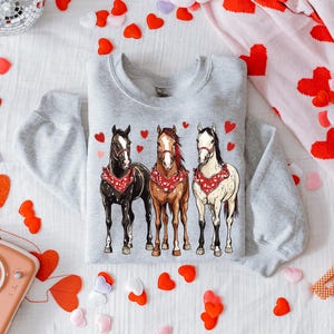 Puede incluir: Una sudadera gris con un gr&aacute;fico de tres caballos con pa&ntilde;uelos. Los caballos son marr&oacute;n, blanco y negro. El fondo es una superficie blanca con corazones rojos.